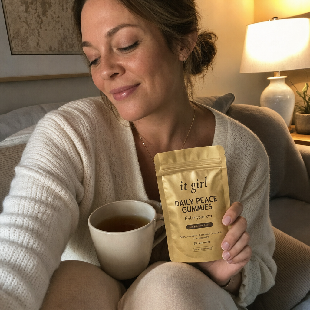 A calm woman curled on her couch in evening lamplight with a mug of tea and the gold It Girl Daily Peace pouch resting in her hand