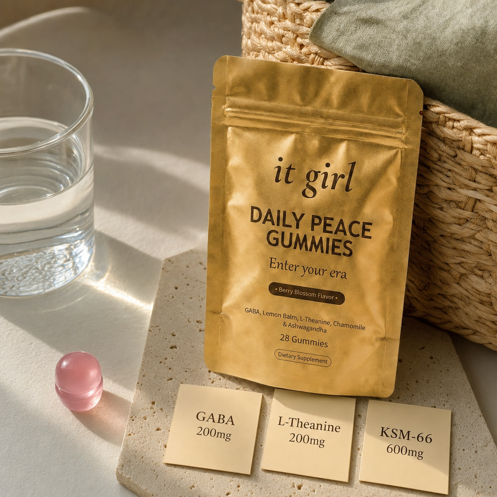 Warm still life of the matte gold It Girl Daily Peace pouch beside a single pink gummy and three dose cards reading GABA 200mg, L-Theanine 200mg and KSM-66 600mg on a cream surface