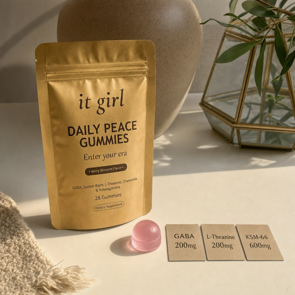 Warm still life of the gold It Girl Daily Peace pouch beside a single pink gummy and three dose cards reading GABA 200mg, L-Theanine 200mg, KSM-66 600mg, the studied stack