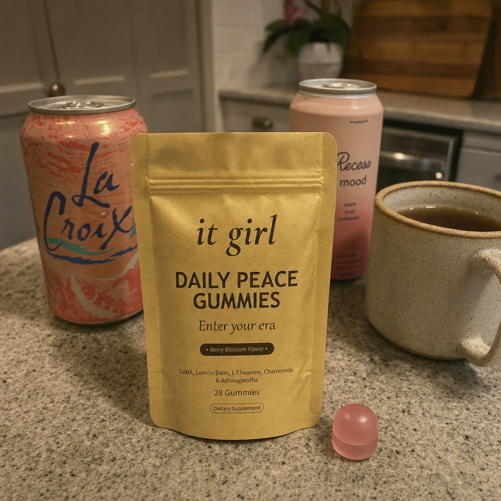 The real matte gold It Girl Daily Peace pouch forward in warm focus on a counter, the abandoned wine swaps, a coral LaCroix can and an adaptogen drink and a mug of cold tea, pushed aside behind it