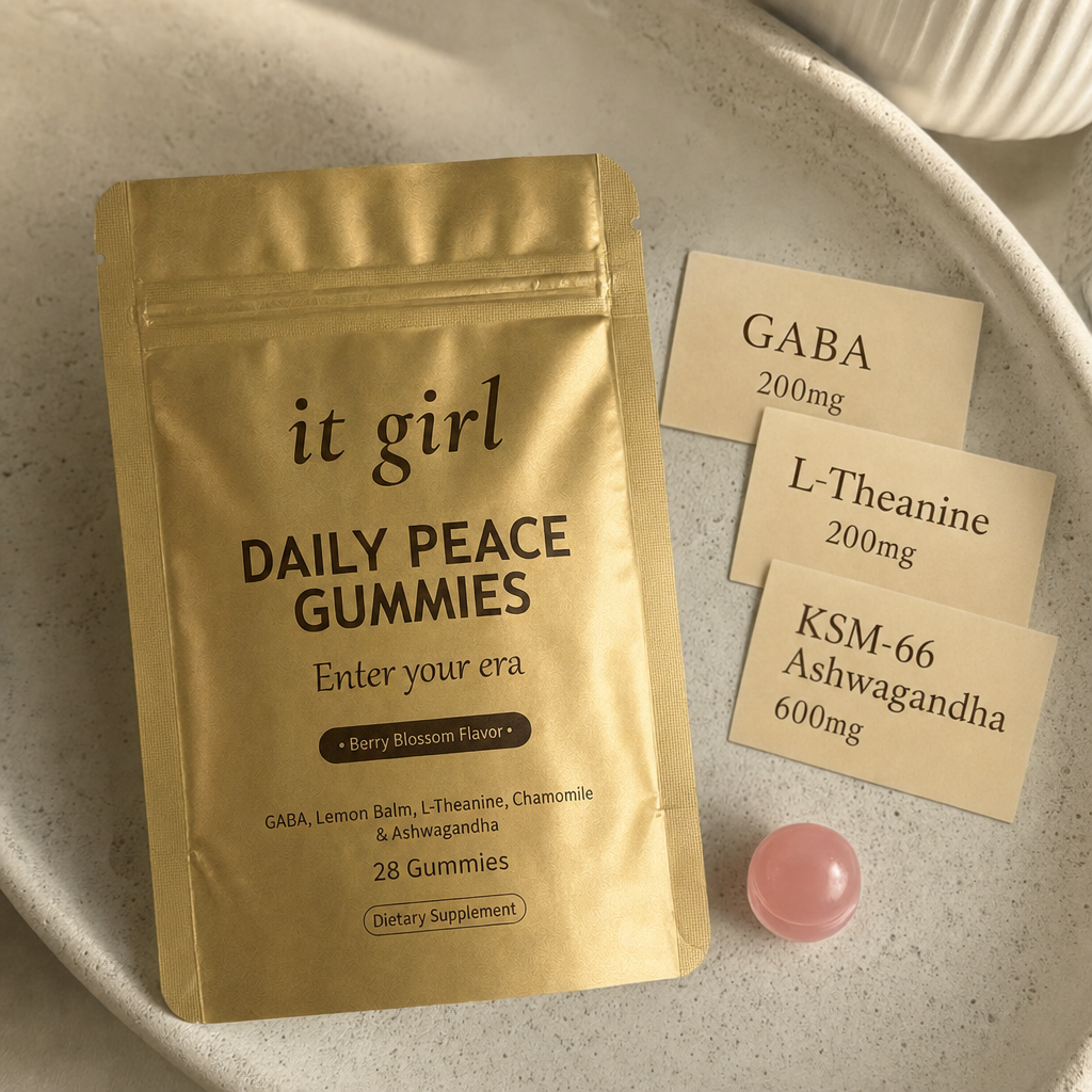 The real matte gold It Girl Daily Peace pouch on warm cream beside three dose cards reading GABA 200mg, L-Theanine 200mg, and KSM-66 Ashwagandha 600mg and a single pink gummy, the formula that hands the calm signal back directly