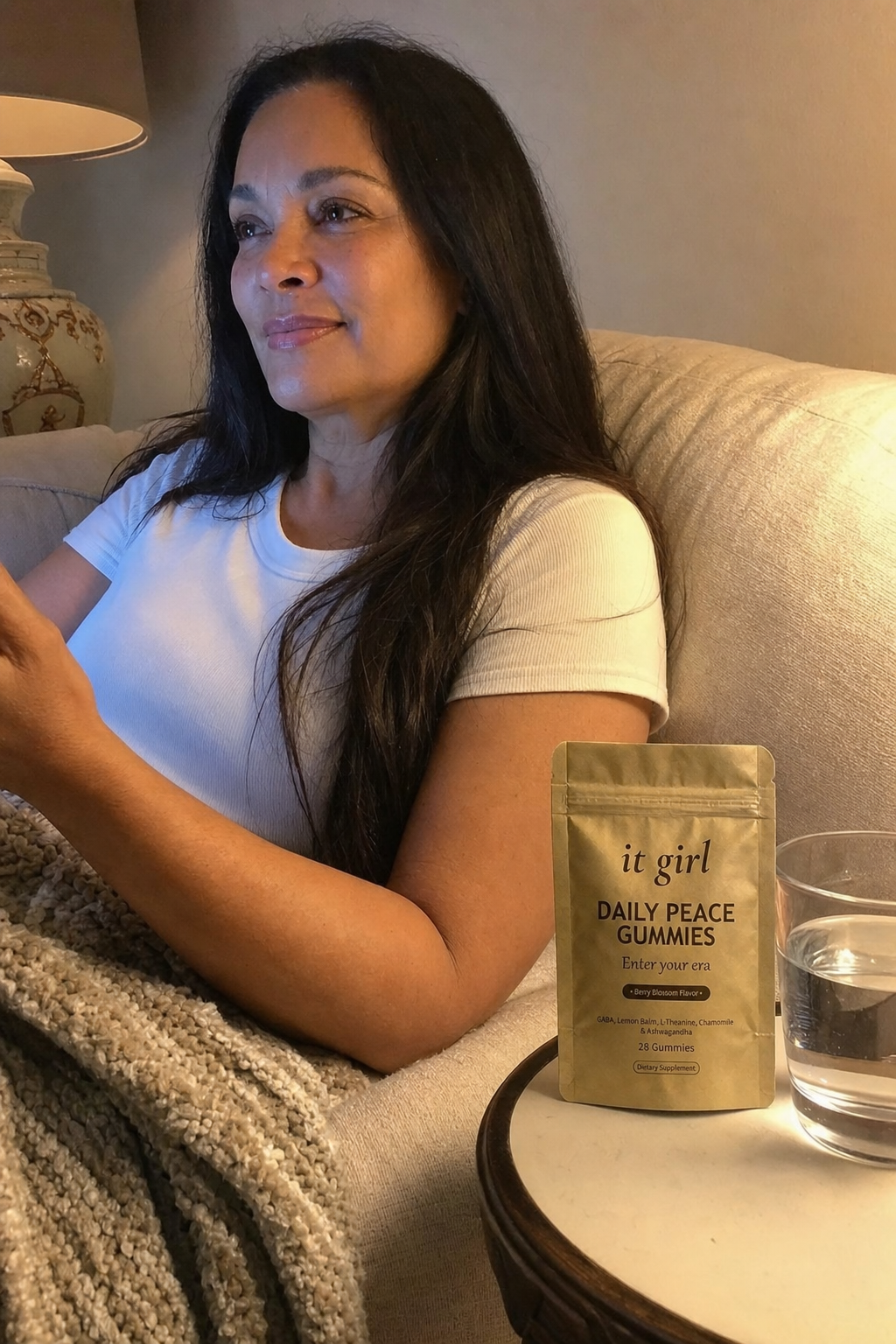 Warm candid evening photo of a woman in her fifties relaxed on her couch at dusk, the gold It Girl Daily Peace pouch and a glass of water on the side table where the wine glass used to be