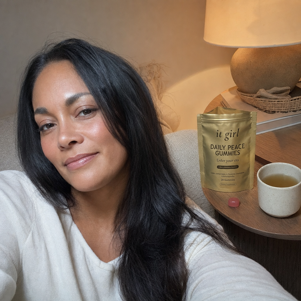 The same calm woman settled on her couch in warm early-evening lamplight with the gold It Girl Daily Peace pouch and a single gummy on the side table, calm not sedation, melatonin-free