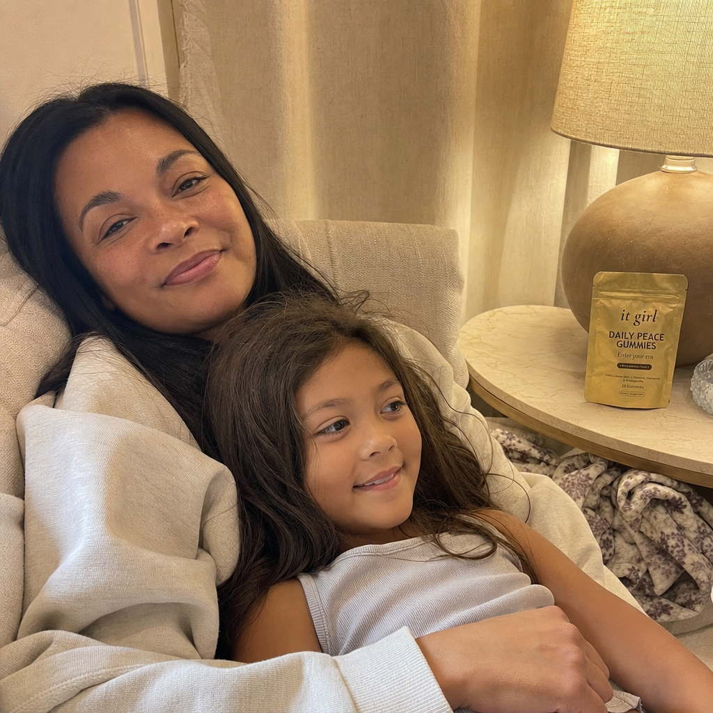 A present mother on the couch in warm lamplight with her young child climbed into her lap and settled in, no bracing, the gold Daily Peace pouch resting in view on the side table