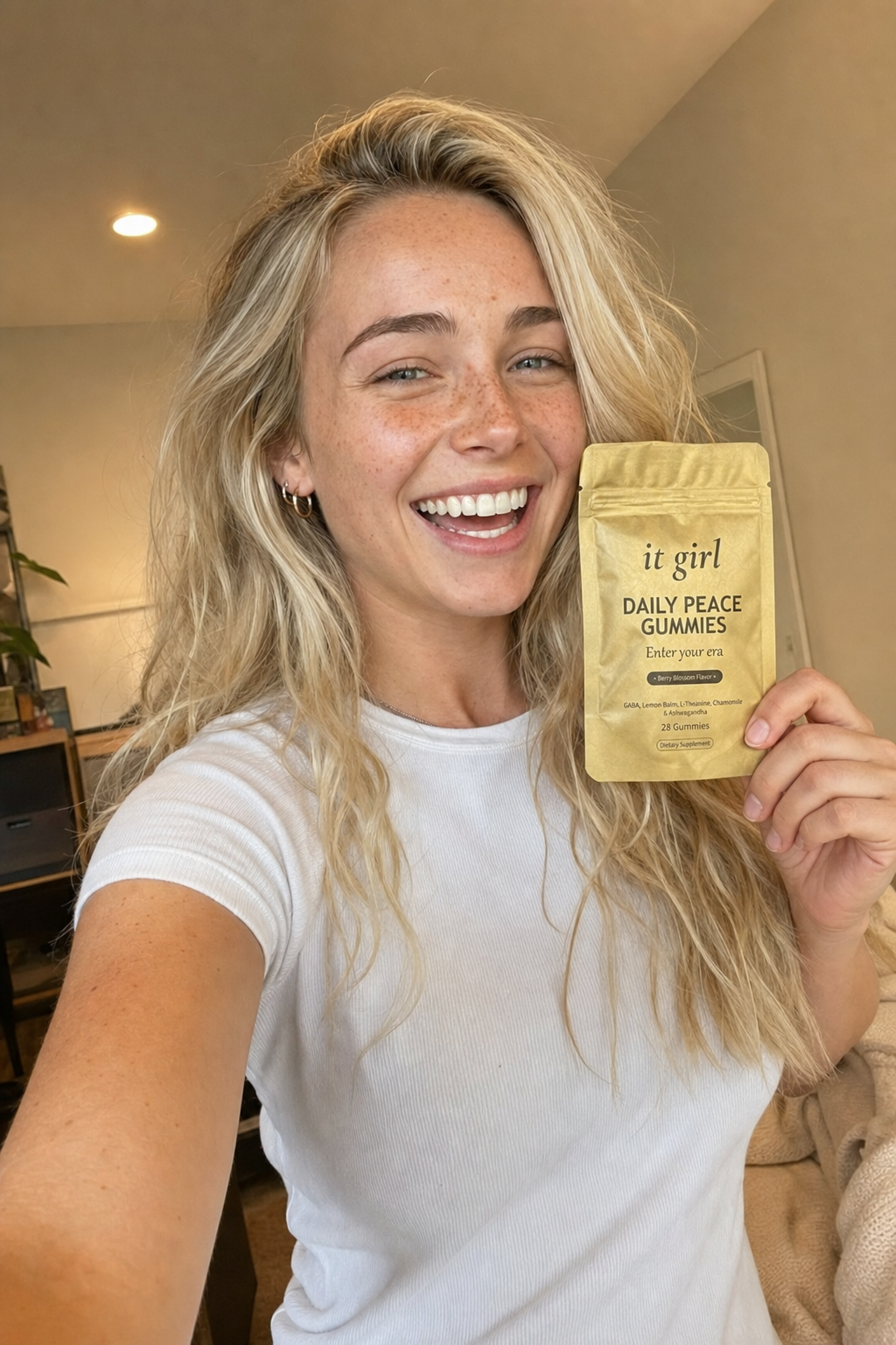 Ultra-realistic candid selfie of Nessa, 24, blonde beach-wave hair and freckles, smiling warmly in soft home light holding the real matte-gold It Girl Daily Peace pouch