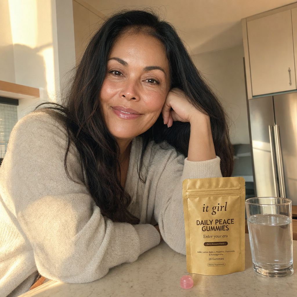 A glowing, restored woman in her early 50s lit from within at a warm kitchen island, her old light back, the gold Daily Peace pouch on the counter and a glass of water, no wine