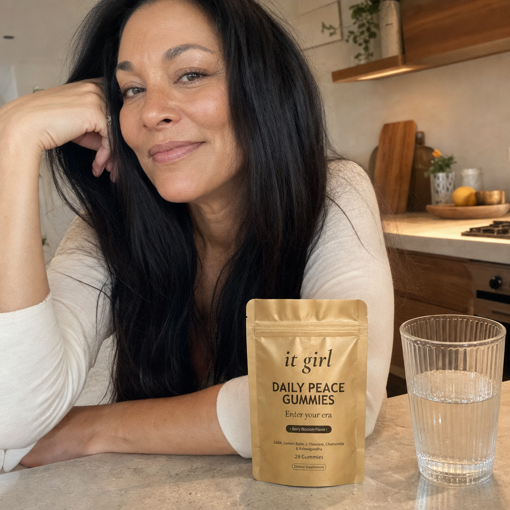 A glowing, restored woman in her early 50s lit from within at a warm kitchen island, her old light back, the gold Daily Peace pouch on the counter and a glass of water, no wine