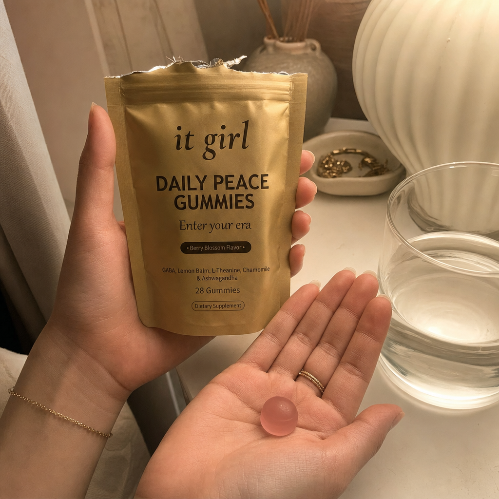 A woman's hand holding the opened matte-gold It Girl Daily Peace pouch with a single pink gummy in warm evening light beside a glass of water on a nightstand, a dollar-a-day evening ritual