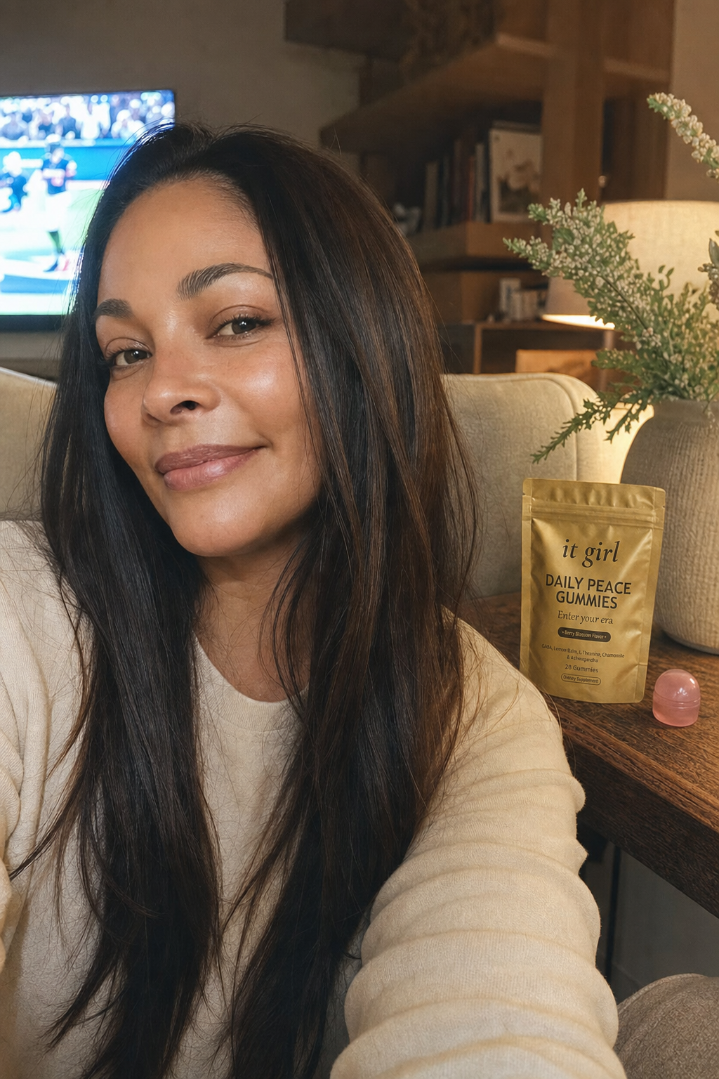 Cristina, a relaxed mom in her late thirties, present and at ease in her warm living room in the evening with the game softly on in the background, the real It Girl Daily Peace pouch on the side table