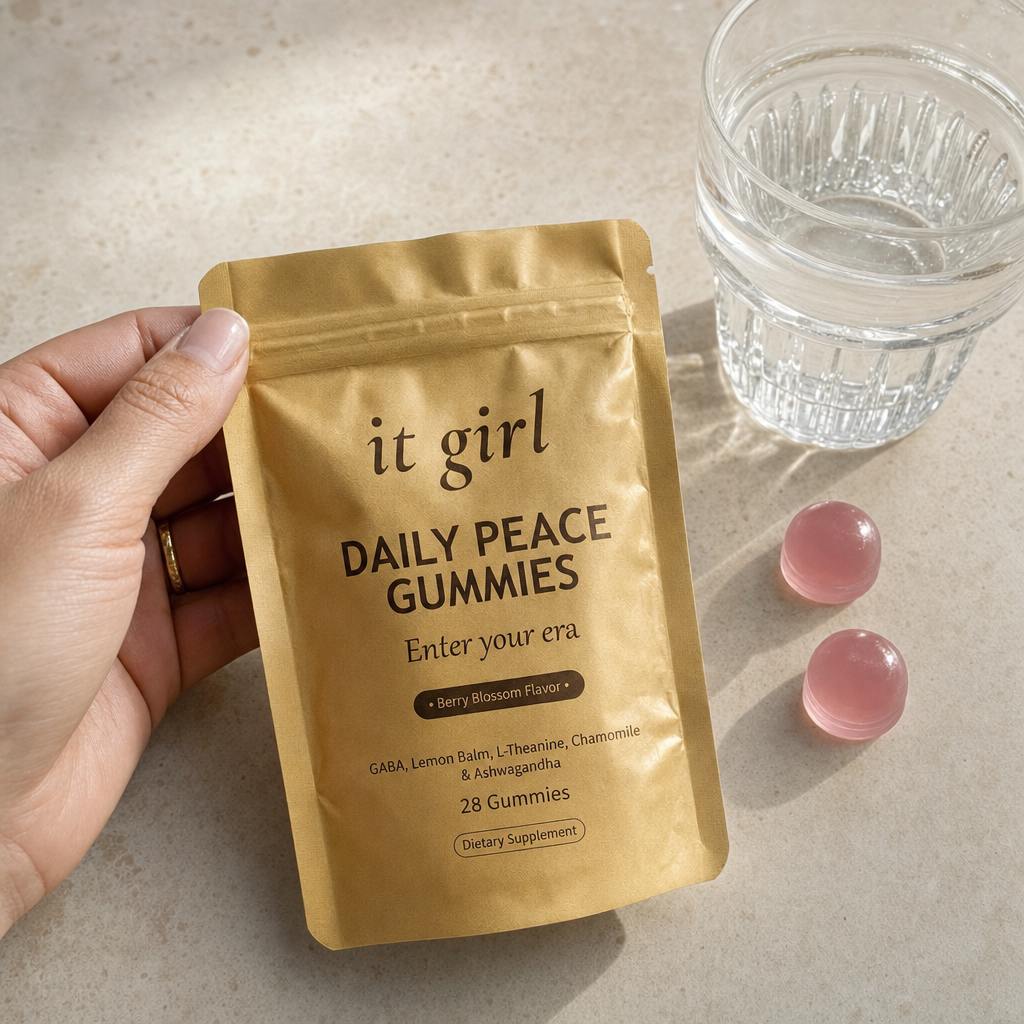 The real soft-gold It Girl Daily Peace pouch laid at palm scale on a warm cream surface with a few gummies and a glass of water, the studied doses in plain daylight