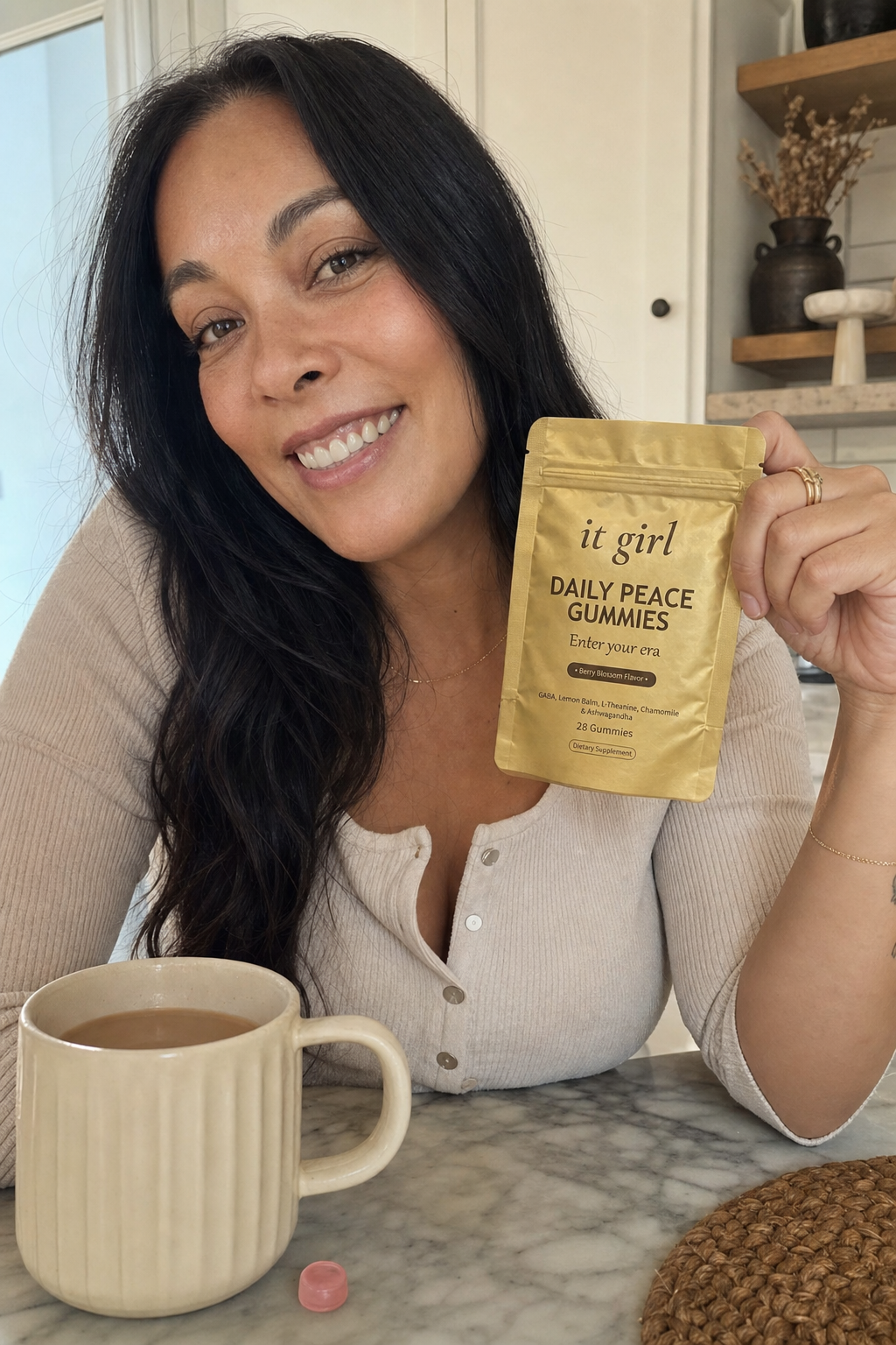 A calm woman in her forties at ease at her morning kitchen counter holding the gold It Girl Daily Peace pouch, settled and in command, coffee nearby
