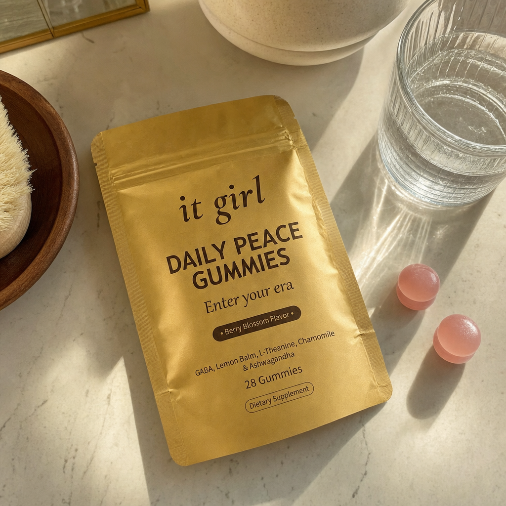 The real gold It Girl Daily Peace pouch laid at palm scale on a warm cream counter with a few gummies and a glass of water, the studied doses in plain daylight
