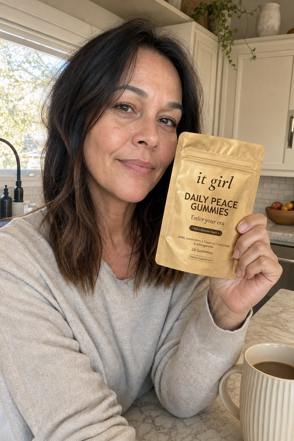 A calm woman in her forties at her morning kitchen counter holding the gold It Girl Daily Peace pouch, settled and in command, coffee nearby