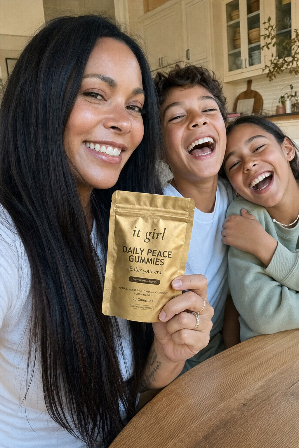 Warm candid arm's-length selfie of a calm, present mom in her forties laughing with her kids at the kitchen table, holding up the gold It Girl Daily Peace pouch, fully in the moment
