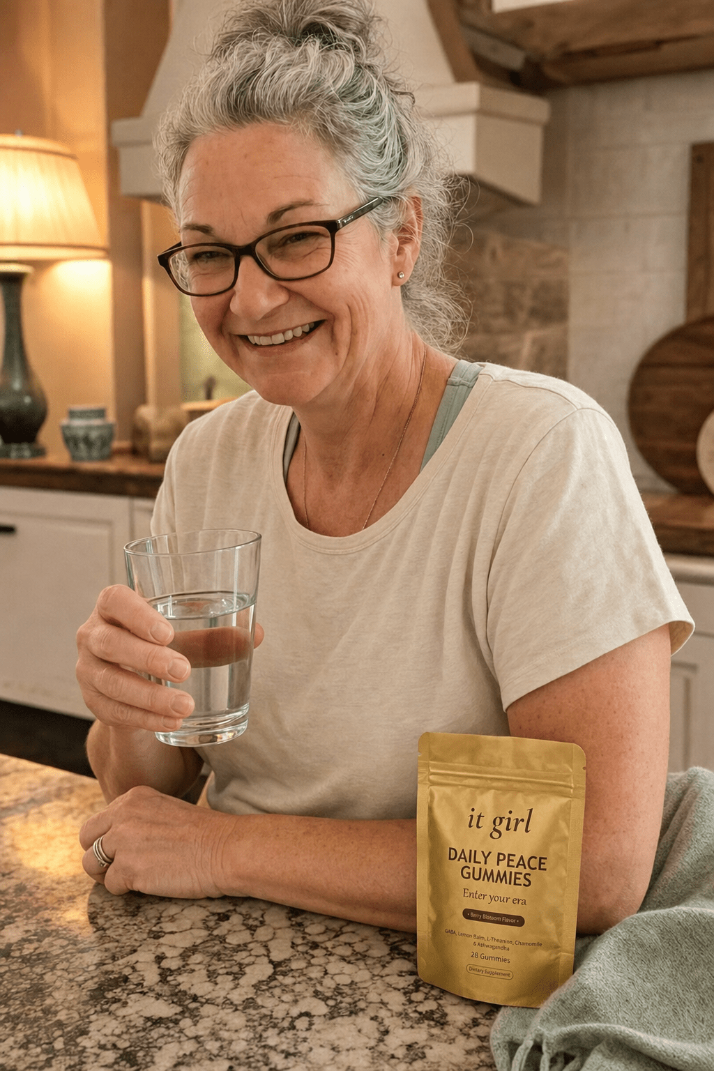 Calm evening candid of a woman about sixty in her warm kitchen, a glass of water in hand and the It Girl Daily Peace pouch on the counter, not reaching for the wine