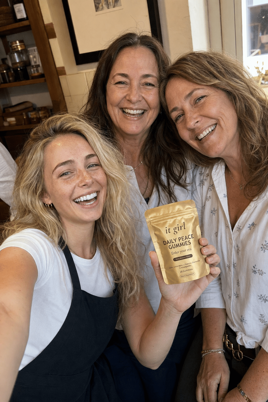 Warm candid arm's-length group selfie of Nessa the young blonde wine-shop cashier and two laughing women regulars in their forties and fifties, holding up the gold It Girl Daily Peace pouch in frame, in a cozy warm-lit setting