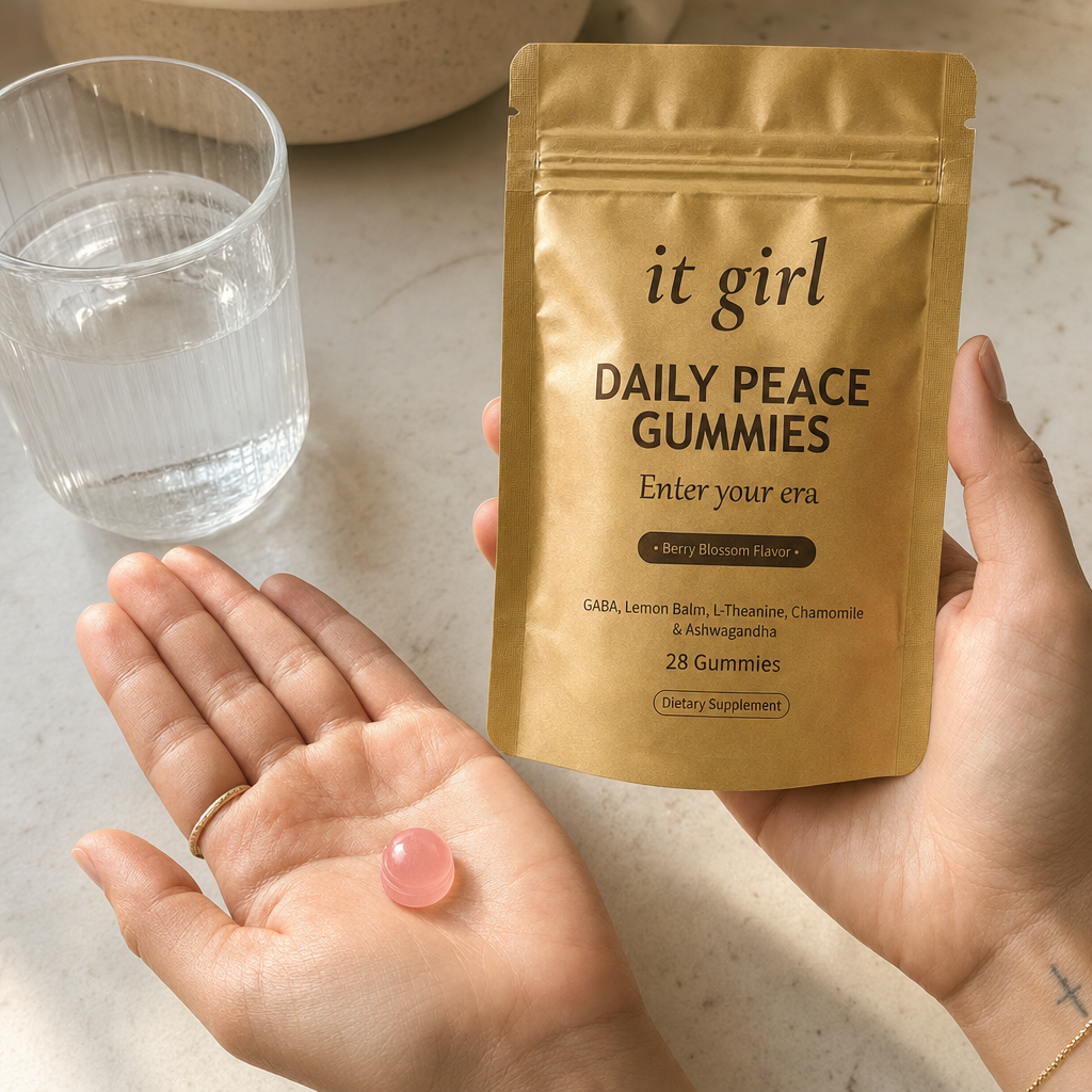 A single pink Daily Peace gummy in an open palm beside the real matte-gold It Girl Daily Peace pouch and a glass of water on a warm cream counter in early-evening light, in full warm color, calm not sedation, the same evening