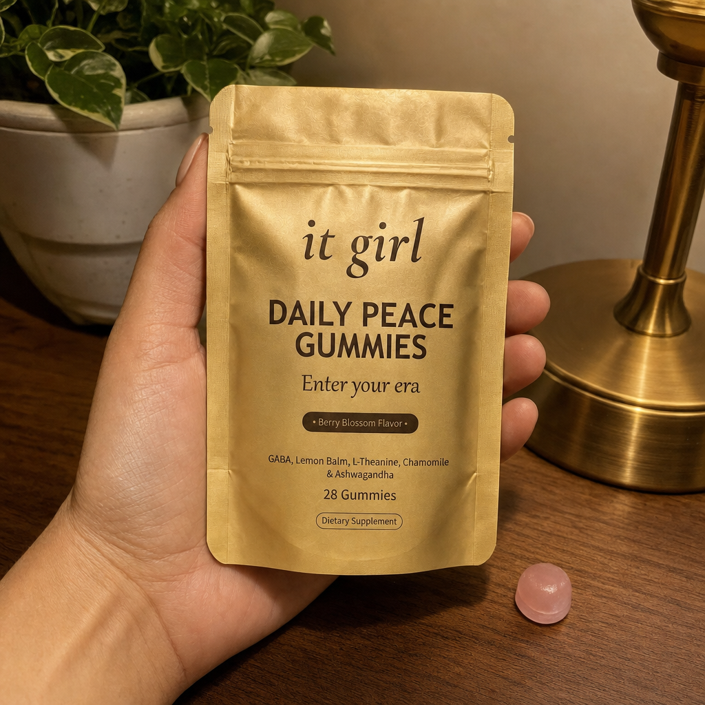 The real matte-gold It Girl Daily Peace pouch resting on Dr. Sofia's warm wood desk in soft evening light beside a single gummy, the vehicle that carries the calm signal clean