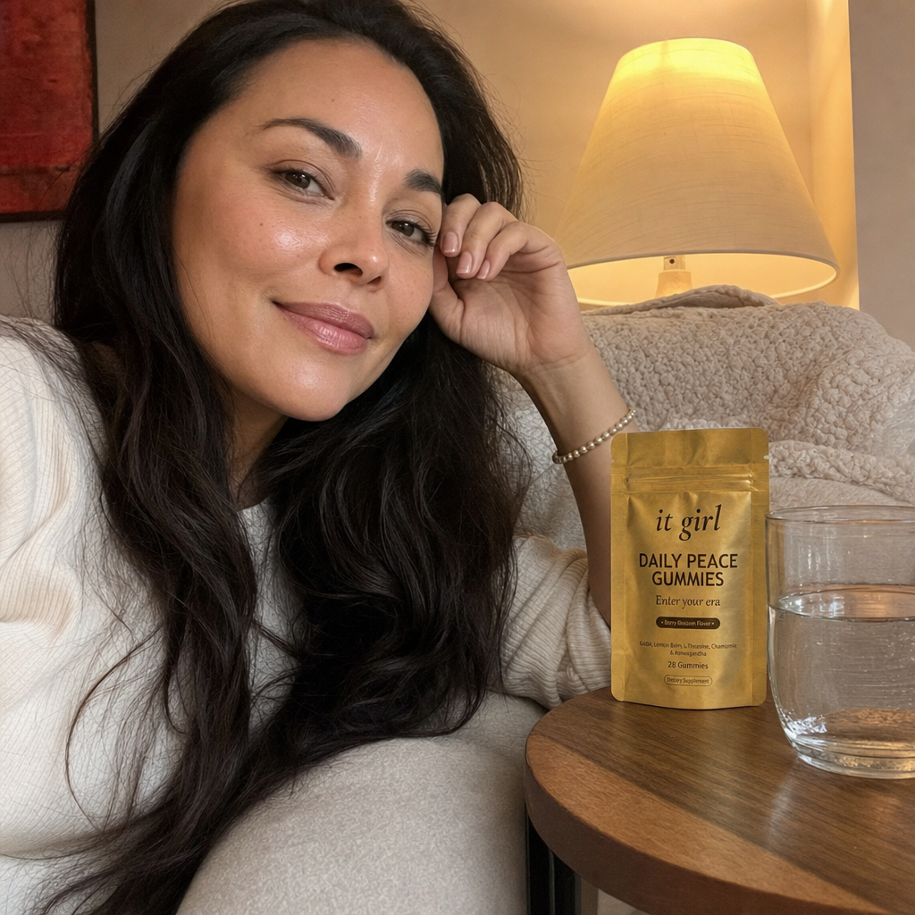 A calm, present woman in her early 40s unwinding in a warm evening living room, the real matte-gold It Girl Daily Peace pouch and a glass of water on the side table, no wine