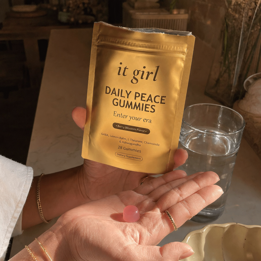 A woman's hand tilting a single pink gummy from the opened real matte-gold It Girl Daily Peace pouch into her palm in warm evening lamp light beside a glass of water