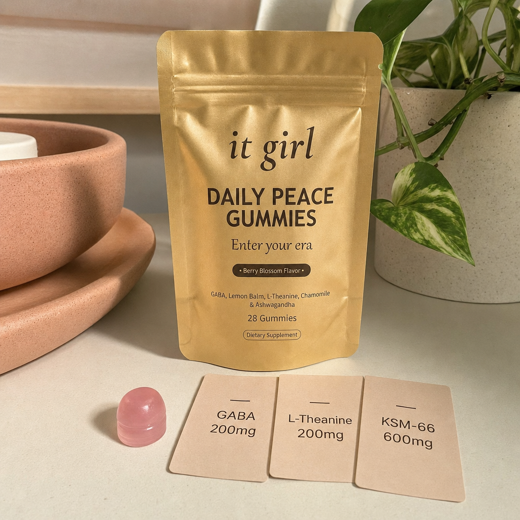 Warm still life on cream: the real matte-gold It Girl Daily Peace pouch with a single pink gummy and three dose cards reading GABA 200mg, L-Theanine 200mg, KSM-66 600mg