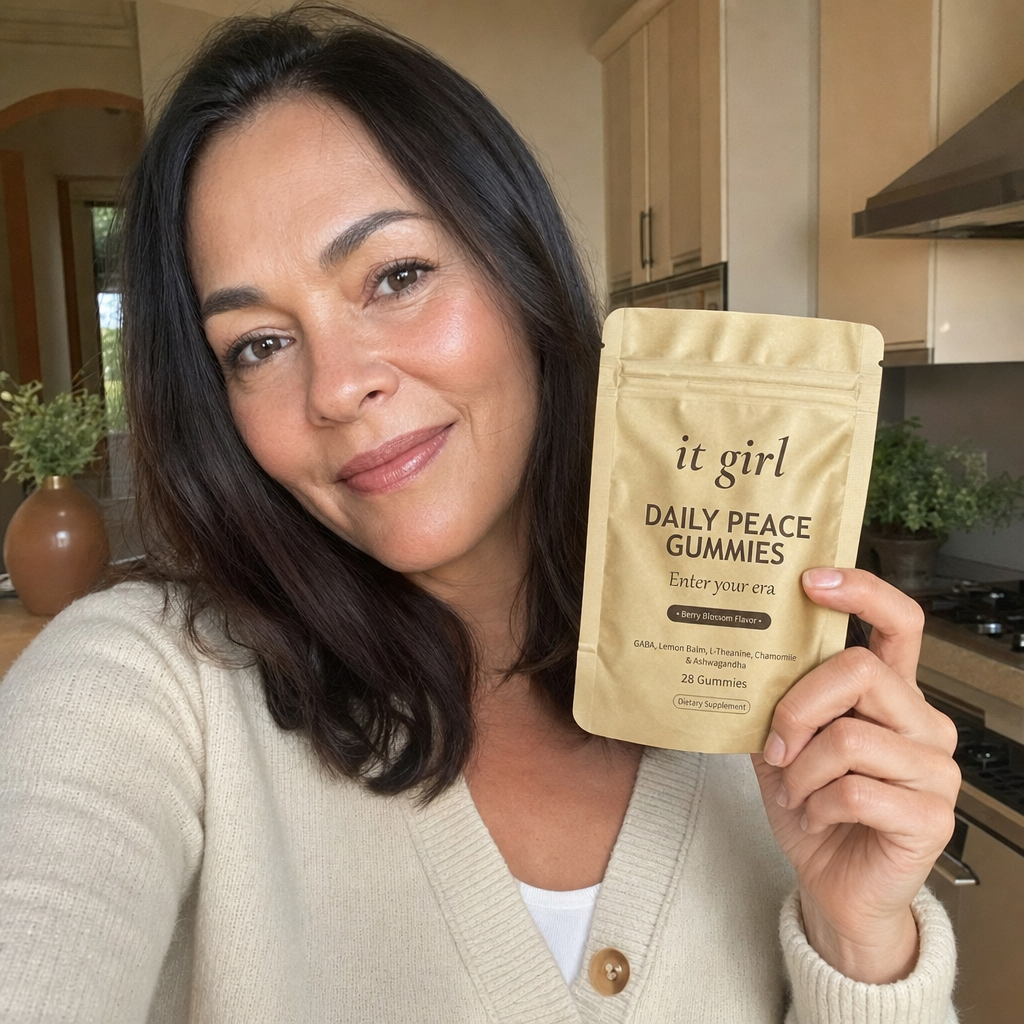 A capable woman in her forties in her warm early-evening kitchen holding the gold It Girl Daily Peace pouch with a relieved calm expression, no wine glass