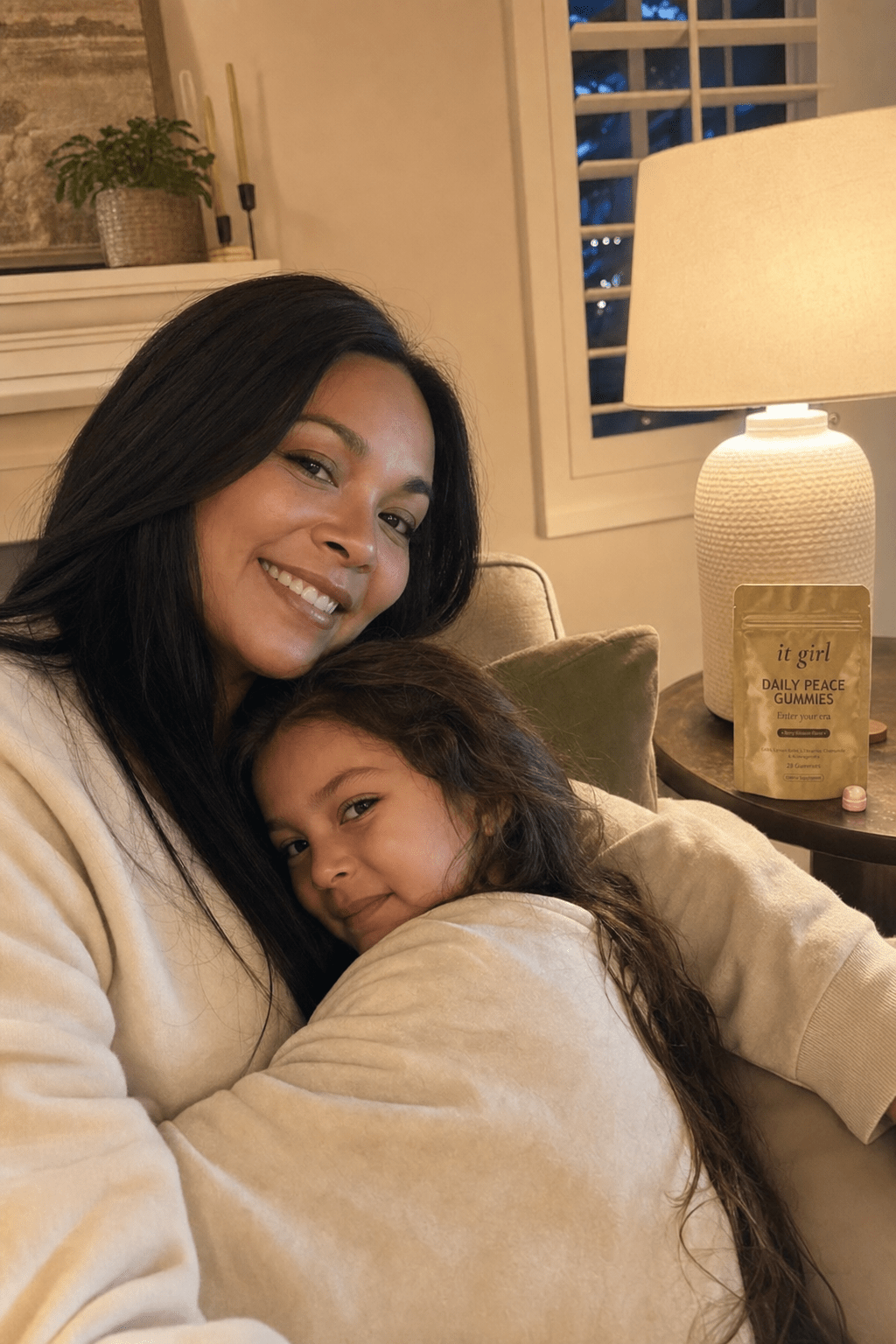 Warm calm evening at home, a composed mother with her child settled easily against her, the matte gold Daily Peace pouch on the side table, the resolved after state