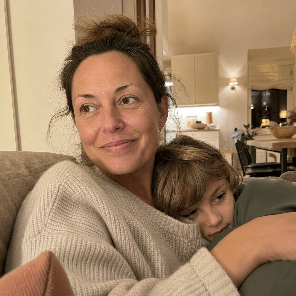 Mother in soft lamp light at home, shoulders dropped and easy, child leaning calmly into her lap, a genuinely calm dinner-table evening