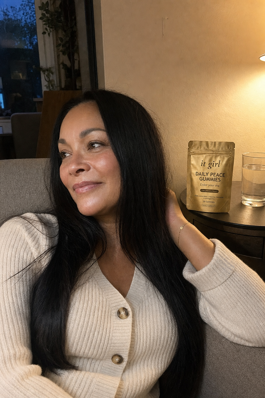 Warm candid evening photo of a woman in her fifties relaxed on her couch at dusk, the gold It Girl Daily Peace pouch and a glass of water on the side table where the wine glass used to be