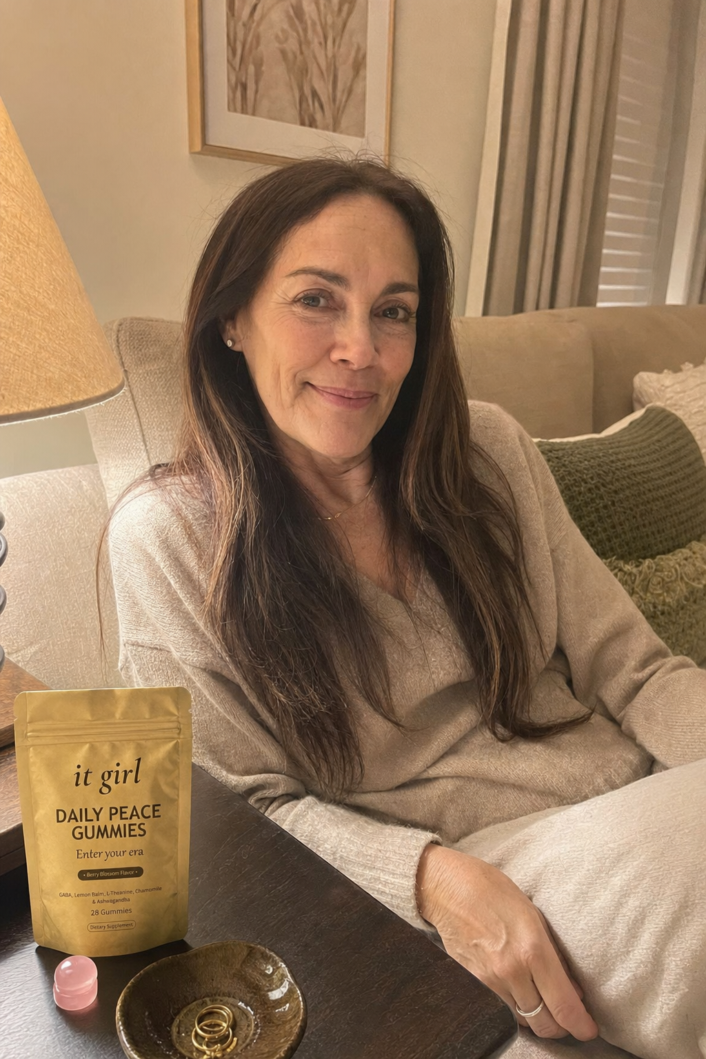A calm woman in her late fifties at ease on her couch on an ordinary evening, laptop closed, the real matte gold It Girl Daily Peace pouch resting on the side table beside her, the reach finally quiet