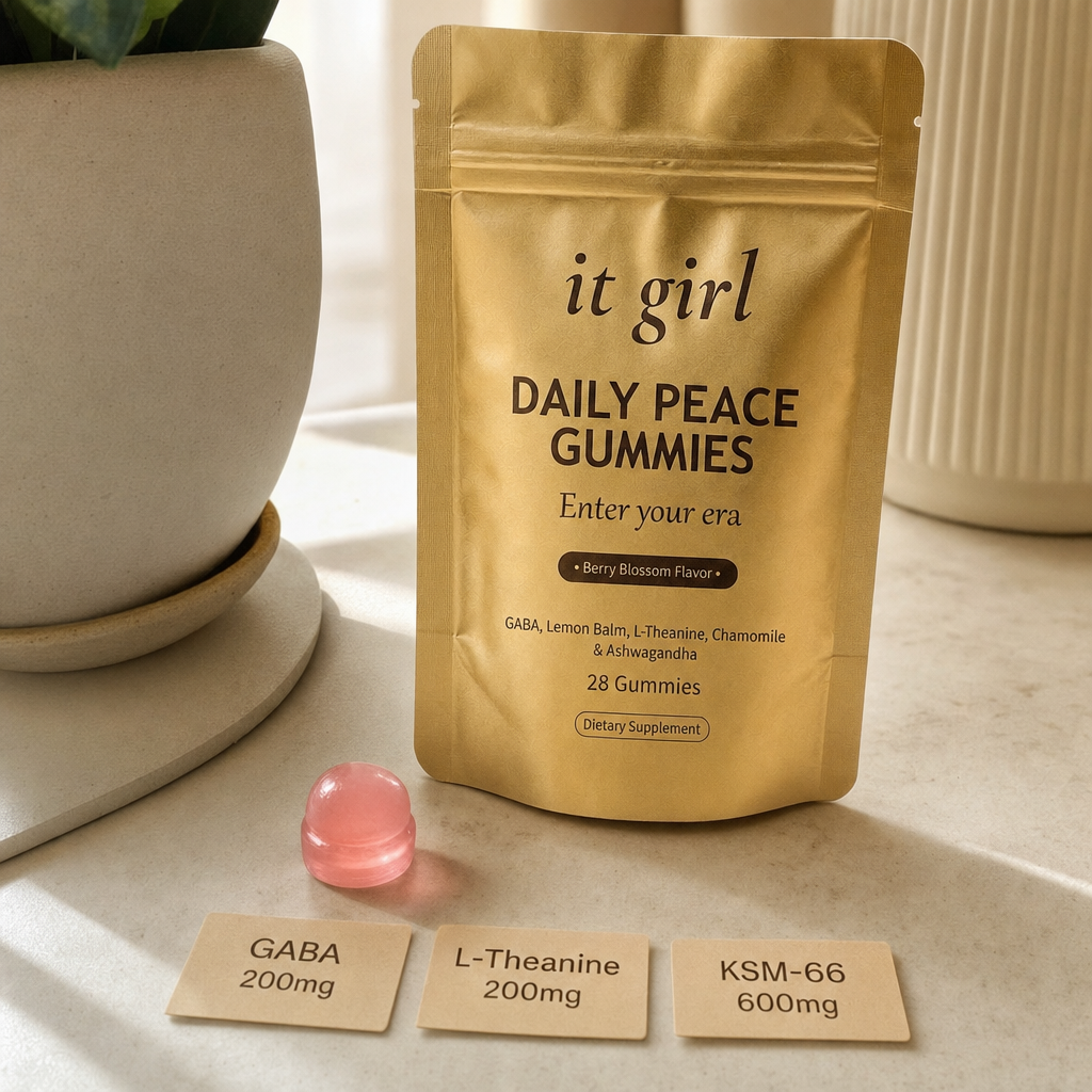 The real matte gold It Girl Daily Peace pouch on a warm cream surface beside a single pink gummy and three named dose cards, GABA 200mg, L-Theanine 200mg, KSM-66 600mg, the studied stack named on the label