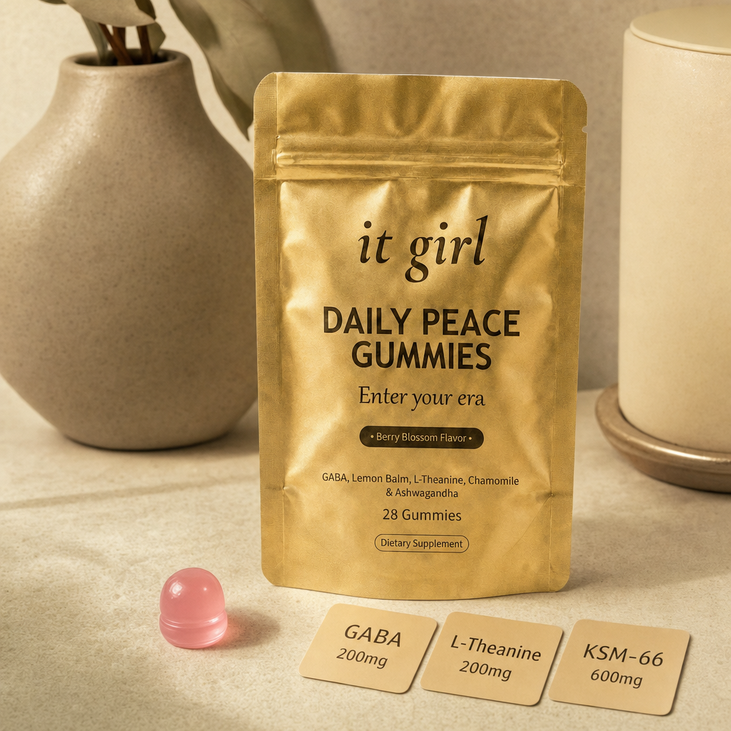 The real matte gold It Girl Daily Peace pouch on a warm cream surface beside a single pink gummy and three named dose cards, GABA 200mg, L-Theanine 200mg, KSM-66 600mg, the studied stack named on the label