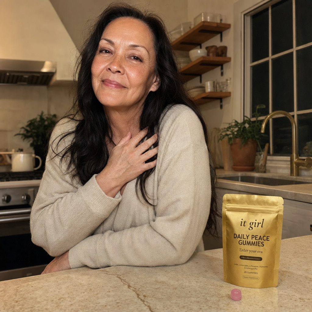 Real woman in her late fifties at ease in a warm-lit evening kitchen, calm and self-forgiving, the gold It Girl Daily Peace pouch resting on the counter, the reach finally understood and no risk in trying