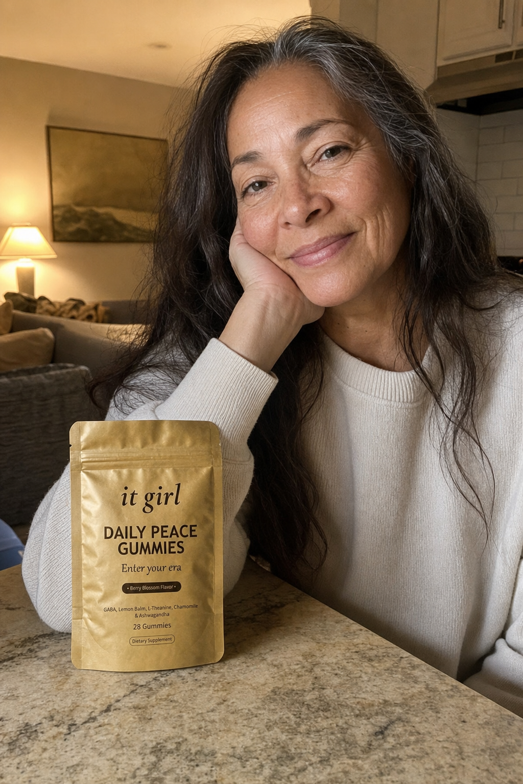 Warm candid evening photo of a relaxed woman in her fifties winding down at home, the real matte-gold It Girl Daily Peace pouch resting on the counter beside her, no wine glass