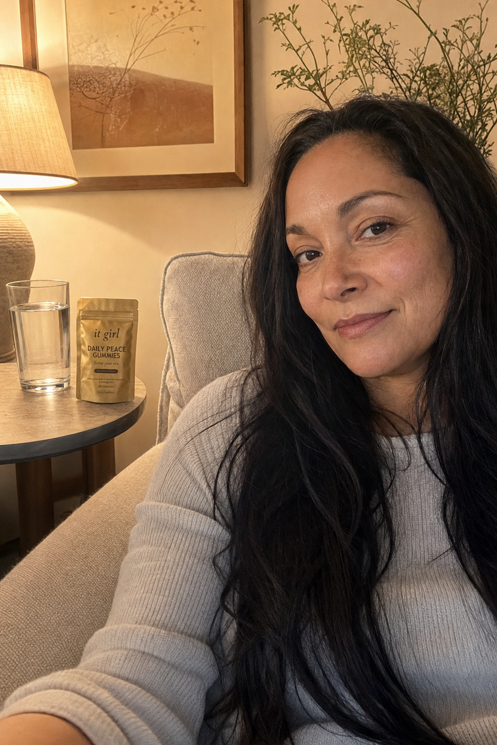 Warm candid evening photo of a woman in her fifties relaxed on her couch at dusk, the gold It Girl Daily Peace pouch and a glass of water on the side table where the wine glass used to be