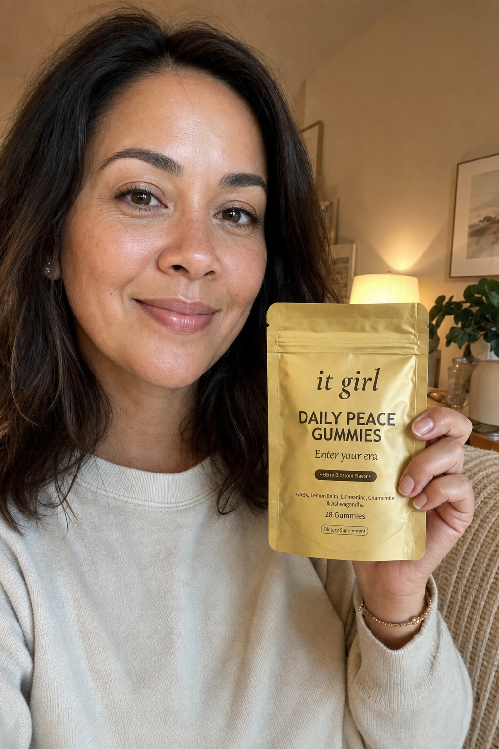 A calm, present mother in her early forties in warm early-evening lamplight holding the real gold It Girl Daily Peace pouch with a relieved half-smile