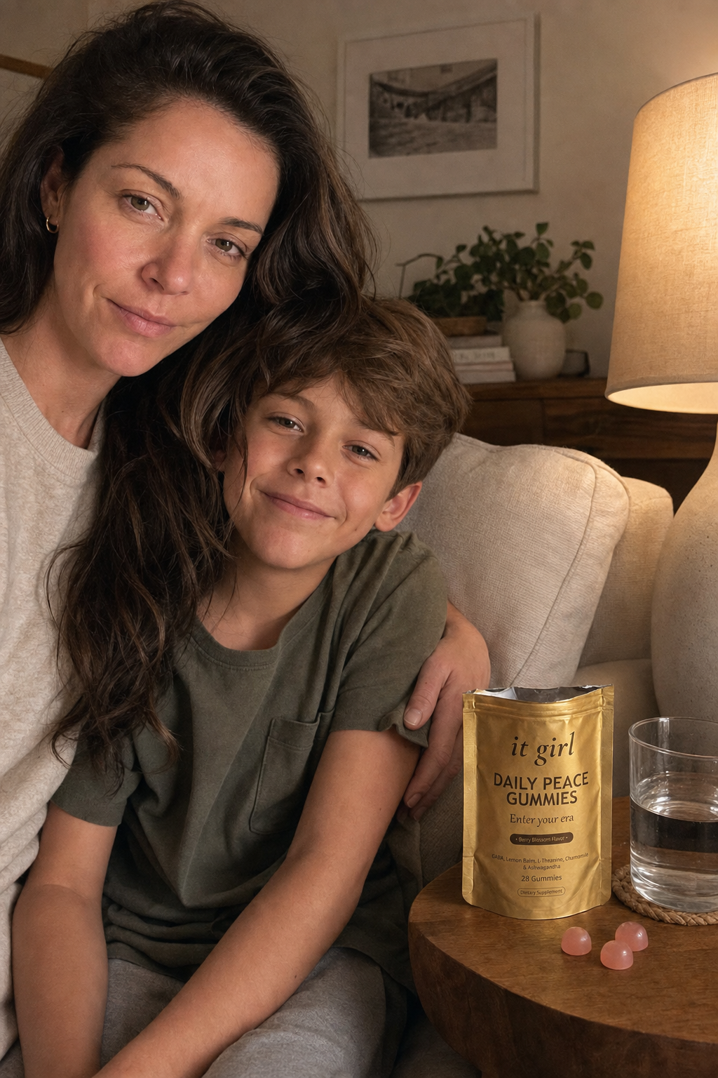 Restored calm mother and her grade-school son sitting close and connected on the couch in warm lamp light, the real palm-sized gold It Girl Daily Peace pouch torn open with a loose pink gummy on the side table beside them