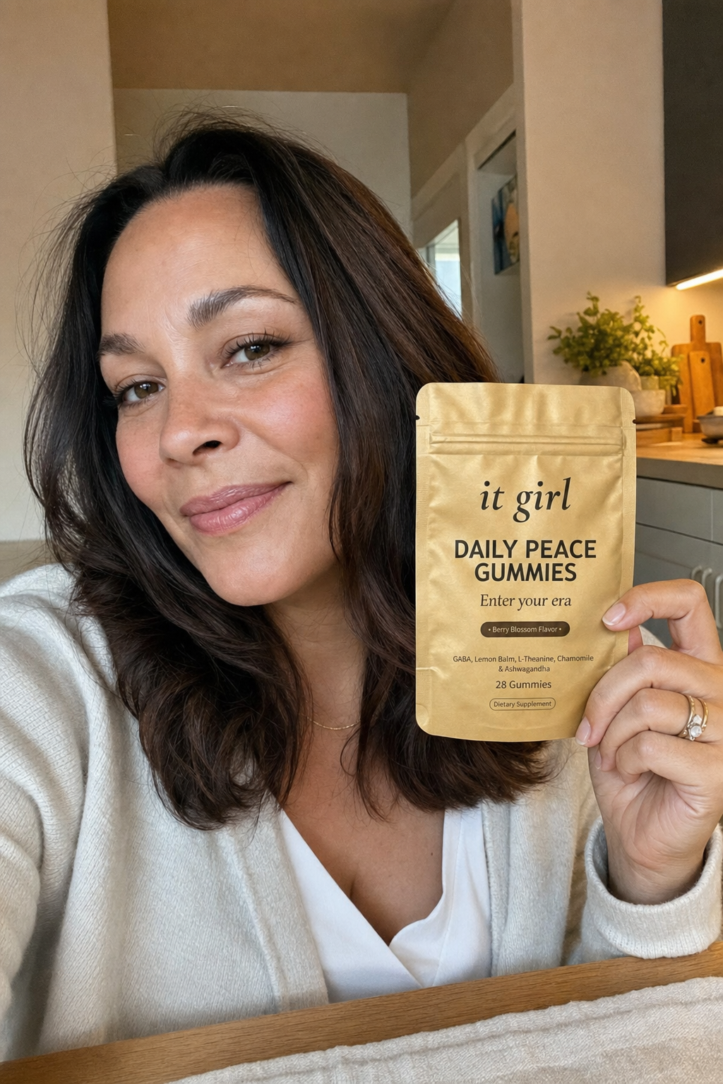 A calm, present woman in her forties at her warm kitchen table holding the real gold It Girl Daily Peace pouch with a relieved half-smile, settled and unbraced