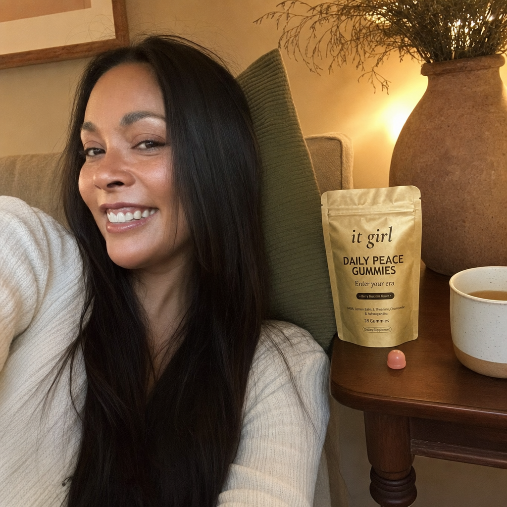The same calm woman settled on her couch in warm early-evening lamplight with the gold It Girl Daily Peace pouch and a single gummy on the side table, calm not sedation, melatonin-free