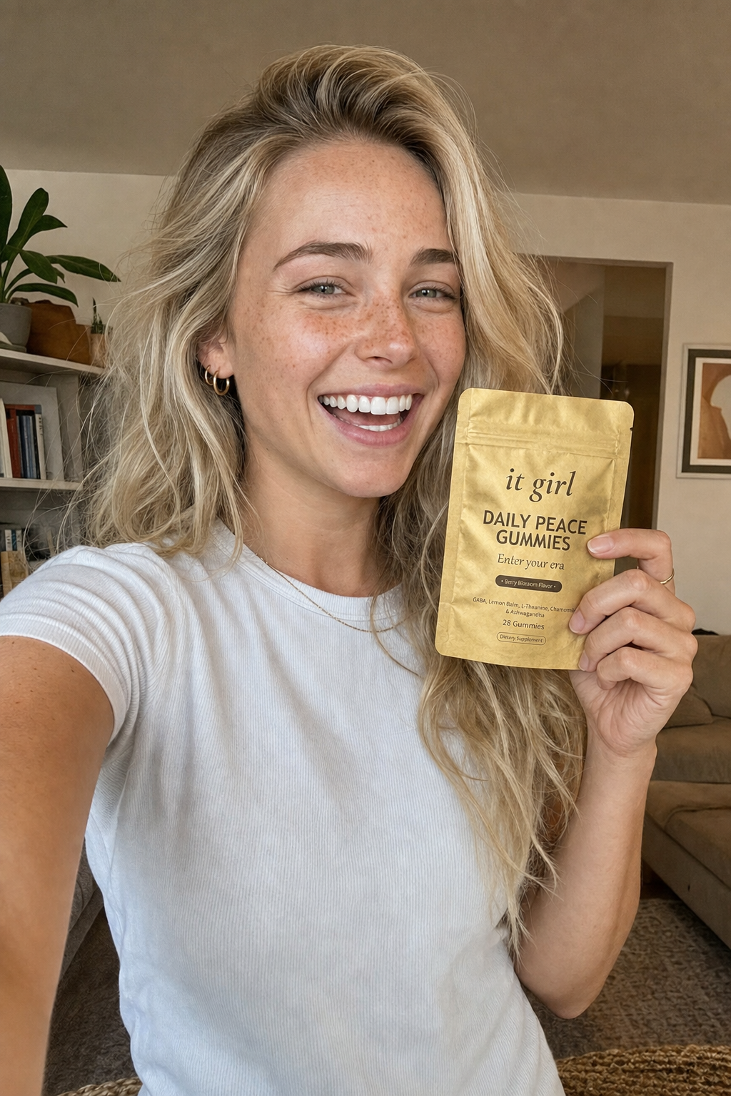 Ultra-realistic candid selfie of Nessa, 24, blonde beach-wave hair and freckles, smiling warmly in soft home light holding the real matte-gold It Girl Daily Peace pouch