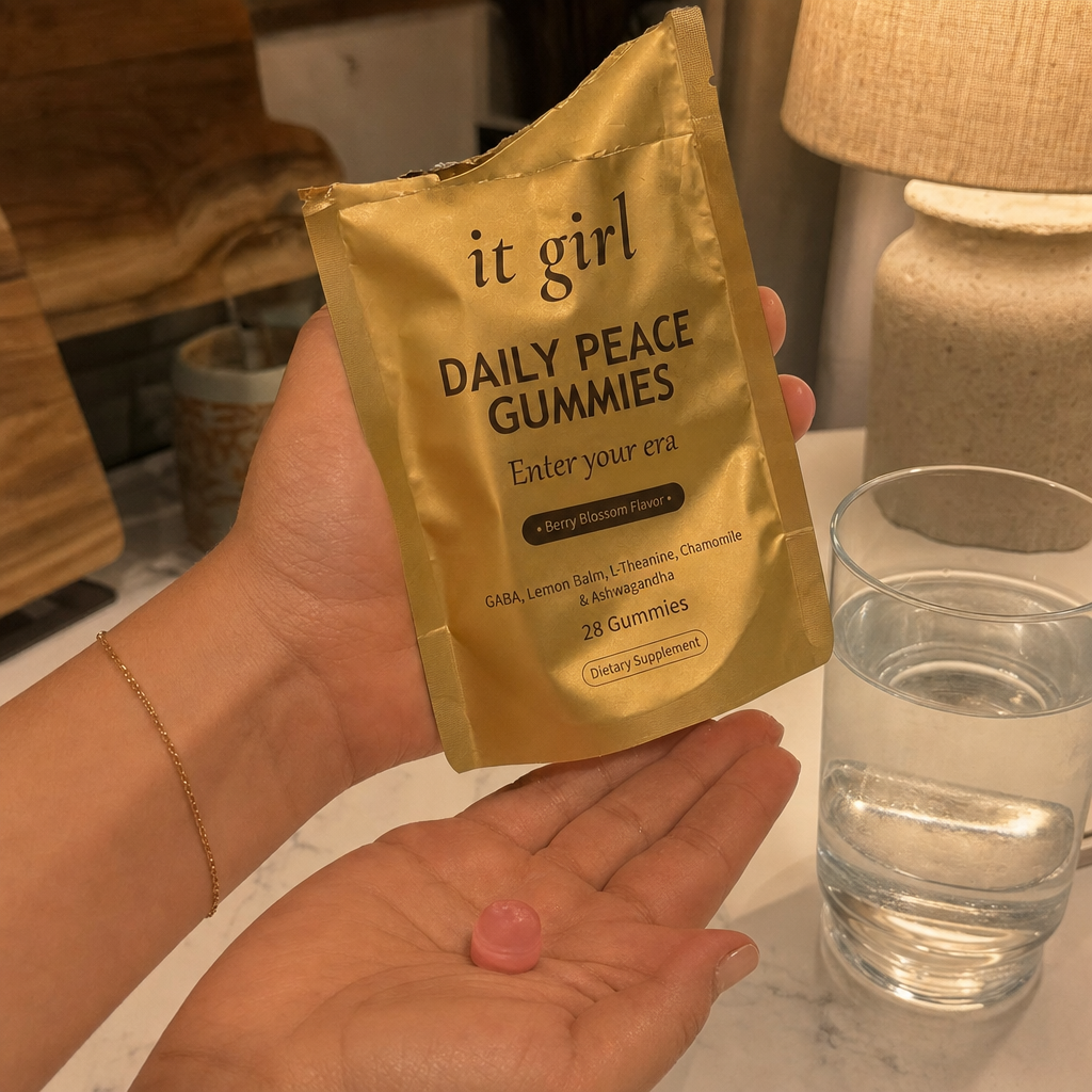 A woman's hand holding the opened matte-gold It Girl Daily Peace pouch tilting a single pink gummy into her palm in warm evening light beside a glass of water, an easy end-of-day ritual