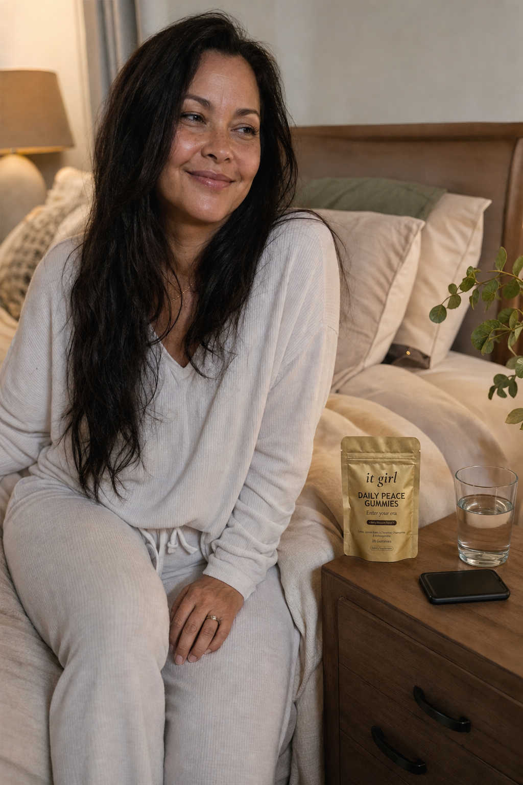 A rested woman on the edge of her bed in warm evening light, phone face-down, the real It Girl Daily Peace pouch on the nightstand beside a glass of water.