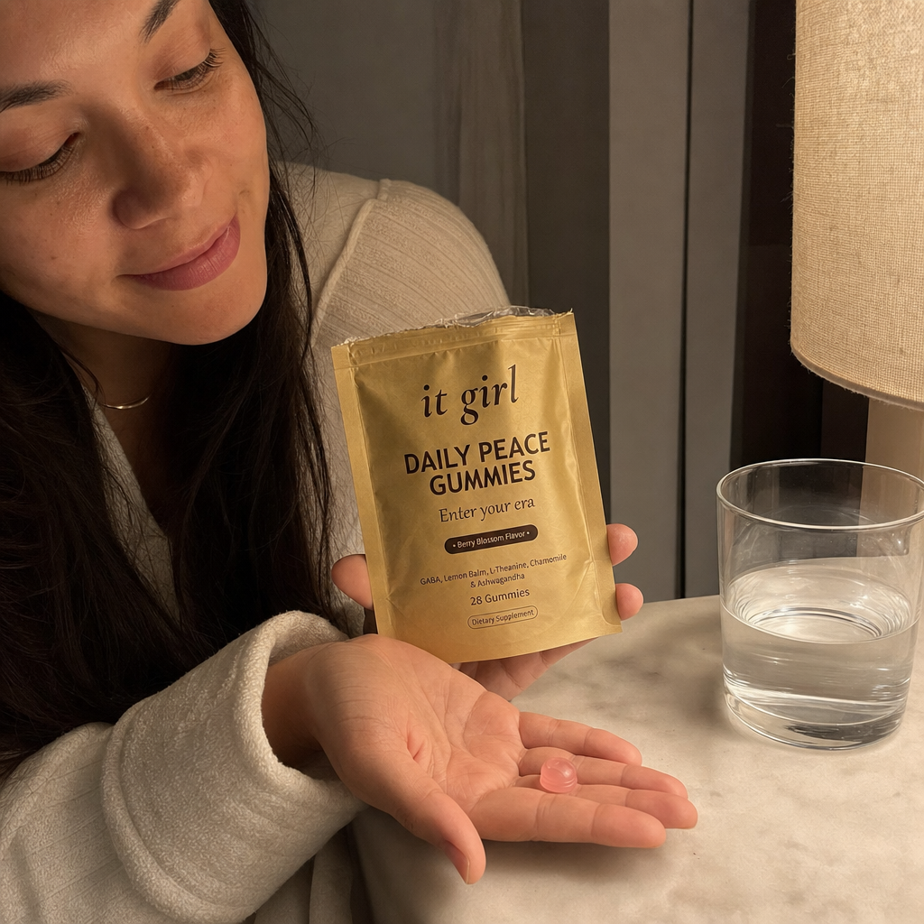 A woman's hand holding the opened matte-gold It Girl Daily Peace pouch with a single pink gummy in warm evening light beside a glass of water on a nightstand, a dollar-a-day evening ritual