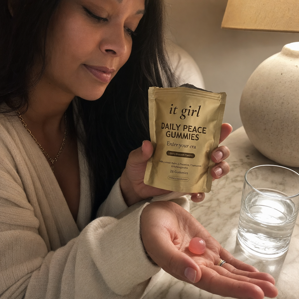 A woman's hand holding the opened matte-gold It Girl Daily Peace pouch with a single pink gummy in warm evening light beside a glass of water on a nightstand, a dollar-a-day evening ritual