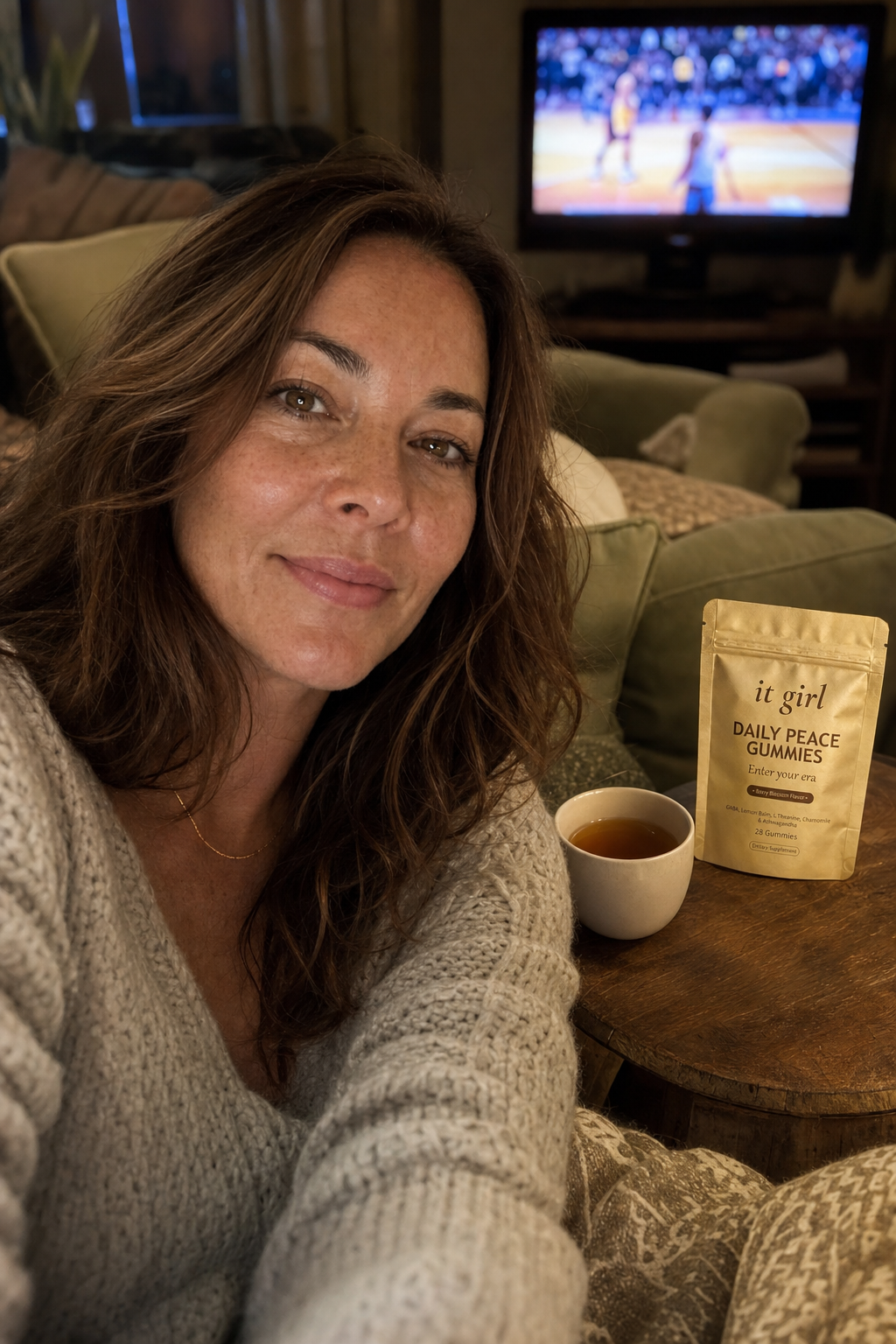Candid evening photo of a calm mom about forty sitting easy on her own living room couch with shoulders down, the game softly glowing on the TV behind her, the gold It Girl Daily Peace pouch on the side table