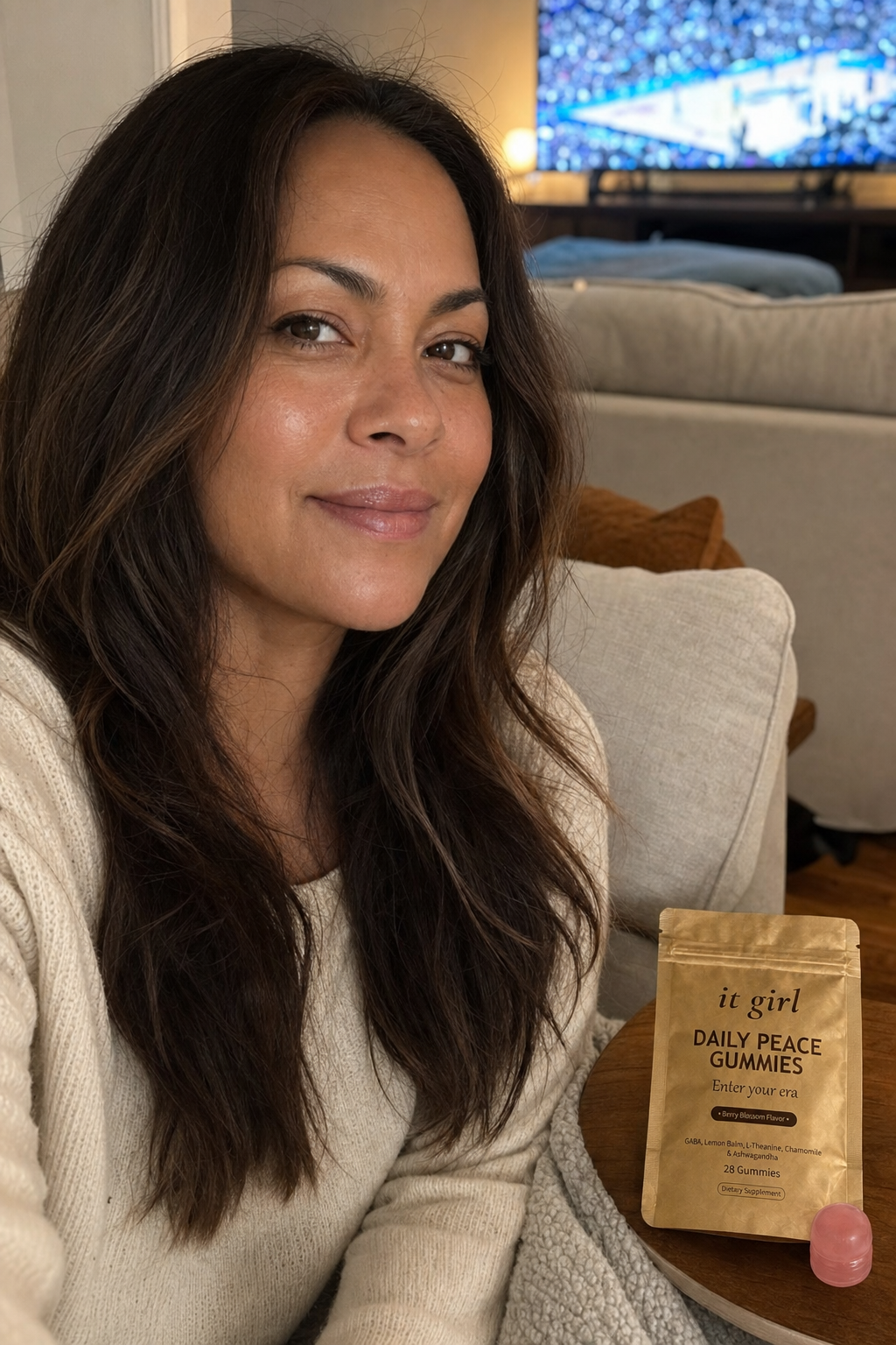 Cristina, a relaxed mom in her late thirties, present and at ease in her warm living room in the evening with the game softly on in the background, the real It Girl Daily Peace pouch on the side table