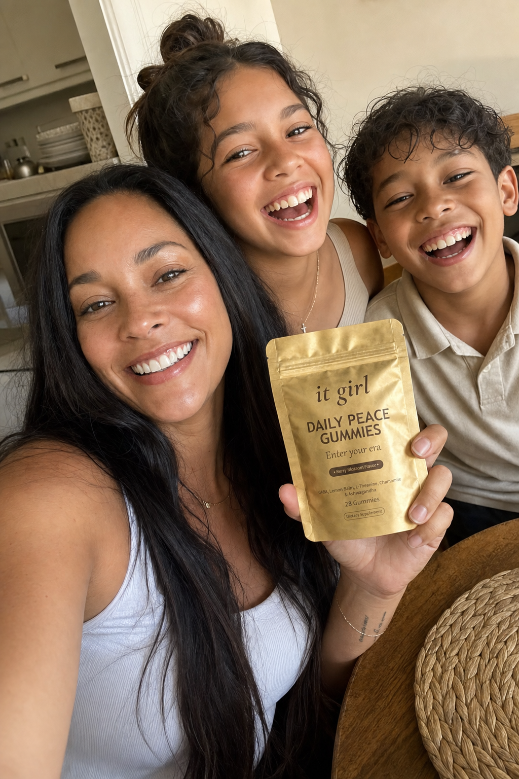 Warm candid arm's-length selfie of a calm, present mom in her forties laughing with her kids at the kitchen table, holding up the gold It Girl Daily Peace pouch, fully in the moment