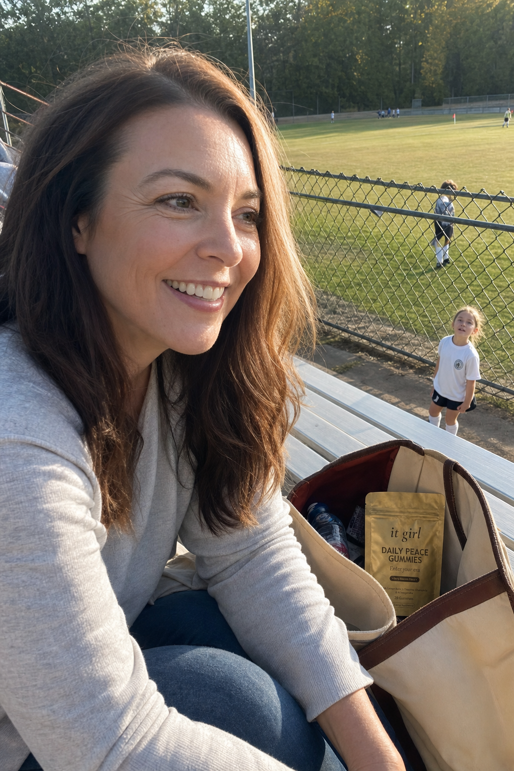 Warm candid moment of a calm, present mother in her late forties up in the bleachers at her kid's soccer game, fully there and smiling toward the field as a small player looks up at her, a gold It Girl Daily Peace pouch resting label-up in her open tote on the bench beside her