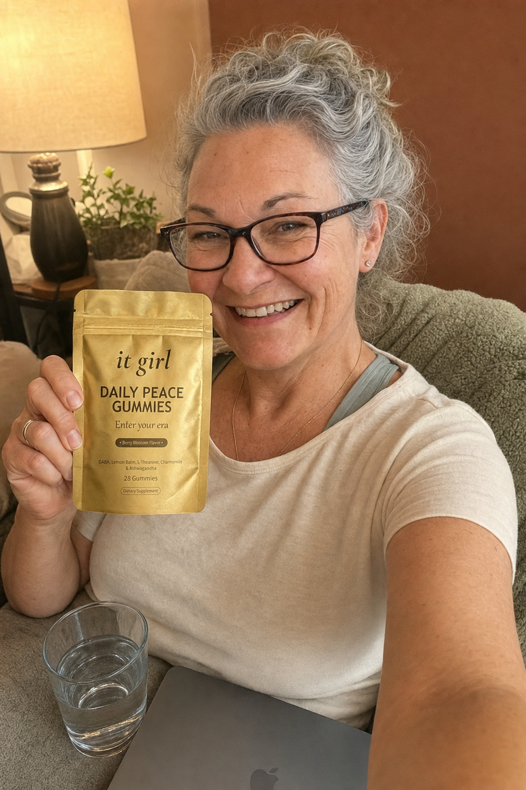 Composed evening selfie of a woman about sixty on her couch at 7:43, laptop closed, holding the gold It Girl Daily Peace pouch, a glass of water nearby, no wine in sight