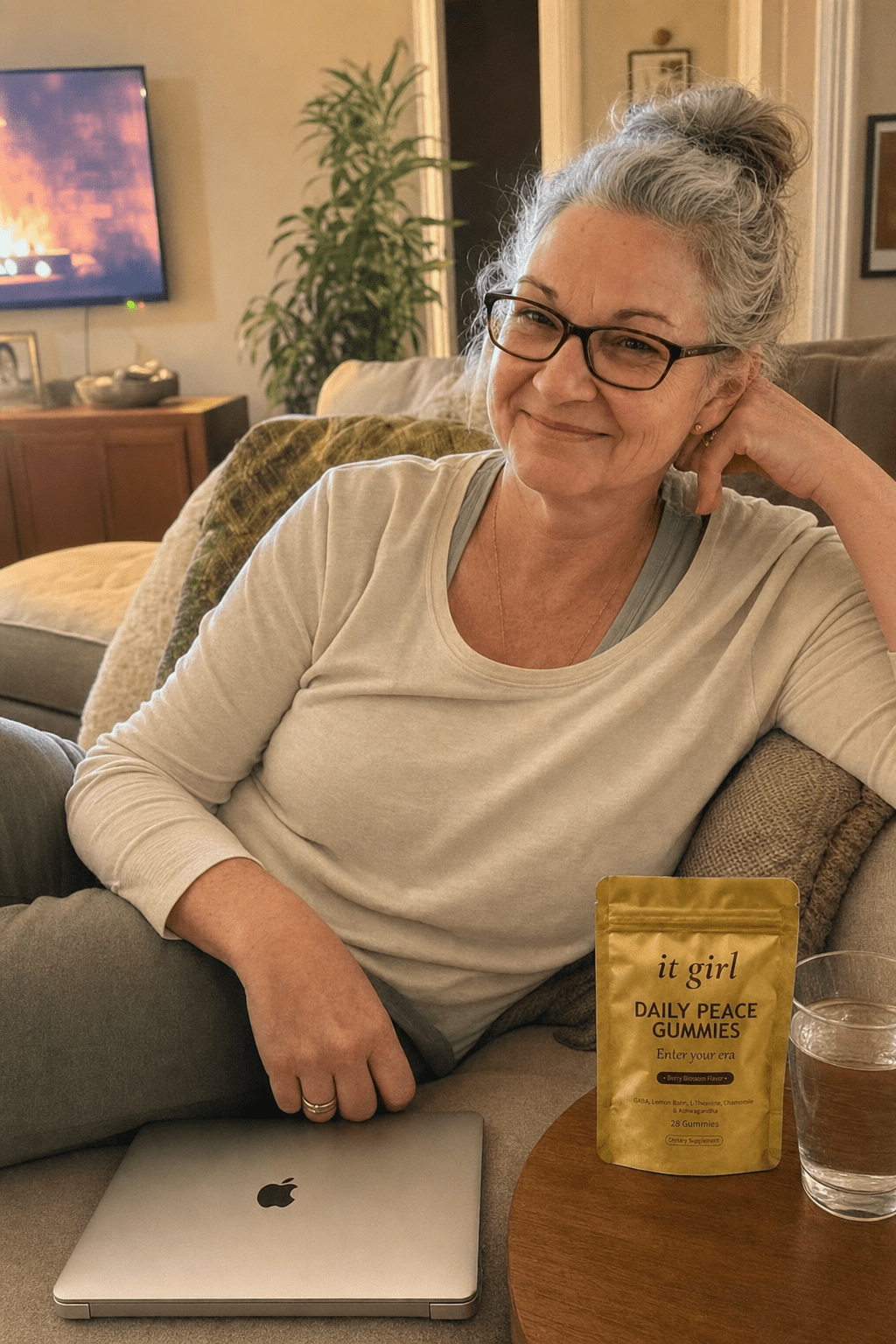 Warm candid evening phone photo of a put-together woman about sixty sitting easy on her own couch, laptop closed, shoulders relaxed, the gold It Girl Daily Peace pouch on the side table beside her and a glass of water, no wine in frame